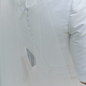 VERONIQUE - Robe de mariée en mikado, le bas s’ouvrant sur des plis creux, ceinture en dentelle rebordée, traîne amovible en organza de soie et petite veste avec boutonnage dans le dos et col fermant avec un noeud. Voile en tulle bordé de dentelle rebrodée.